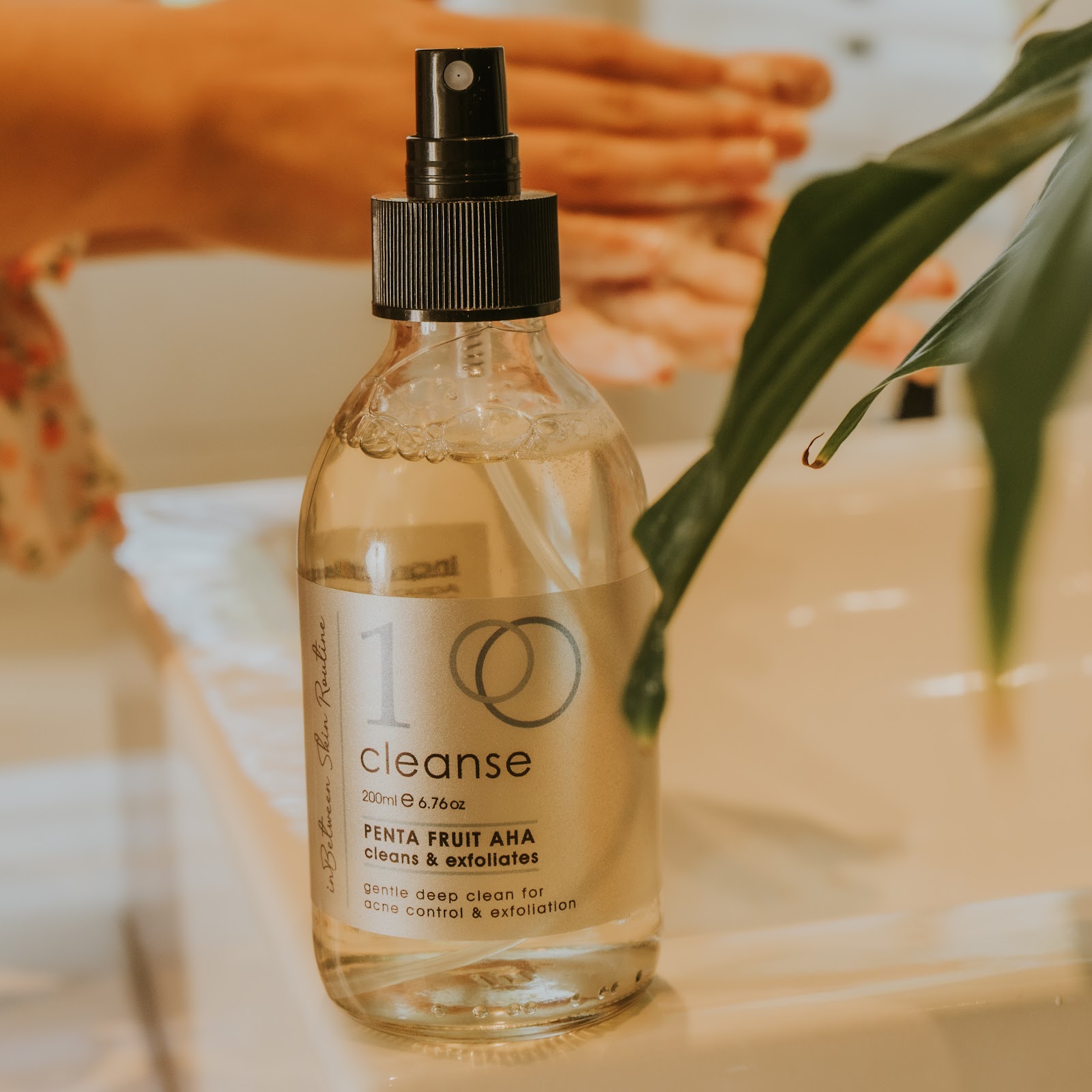 Hands massaging In-Between cleanser beside the cleanse bottle and a fresh leaf